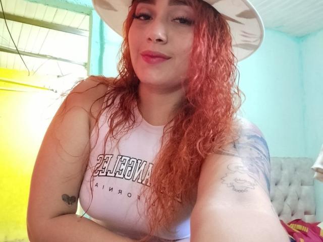 MerydaBraveI with red curly hair, white hat, pink lipstick, white tank top, tattoo on right arm, sitting, smiling, showing tits, light blue wall, yellow and green background.