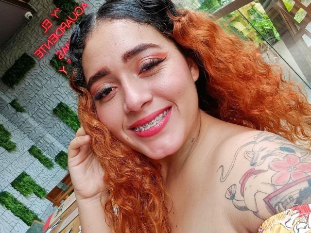 MerydaBraveI has curly orange hair, a smile showing metal teeth, pink eyeshadow, a tattoo on her right shoulder, and is topless with her tits partially visible.