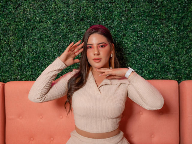EmmaMack with pink eye makeup, wearing a beige ribbed top and skirt, hands near face, sitting on a pink cushioned bench, green wall in background, brown hair with red highlights.