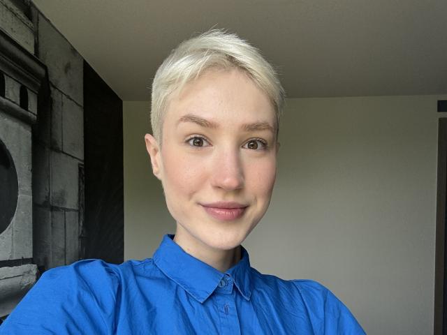 Rhoksi has short, platinum blonde hair, light skin, and brown eyes, wearing a blue shirt, with a subtle smile, showing their nose and lips.