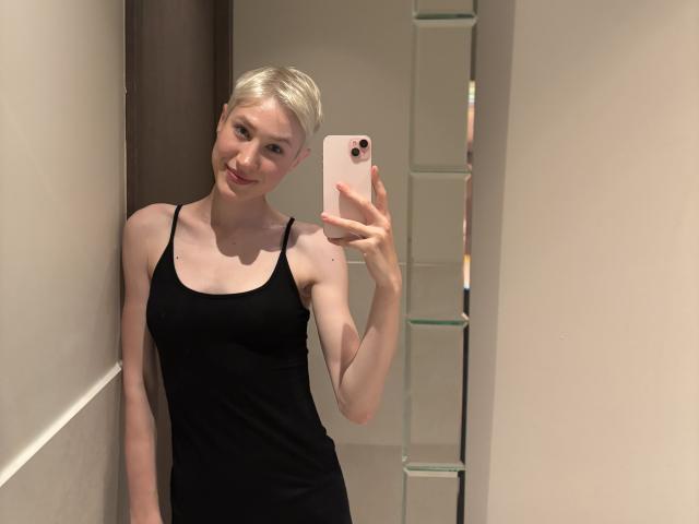 Rhoksi with short blonde hair, wearing a black tank top, holding a pink phone, taking a mirror selfie, smiling, showing a slim, small-ass figure, and pale skin.