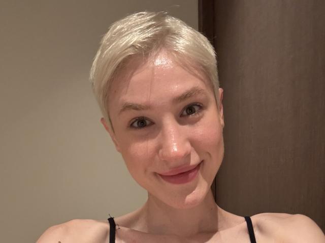 Rhoksi has short, shiny blonde hair, a small nose, and a slight smile. She's wearing a black spaghetti strap top, with her ass barely visible.