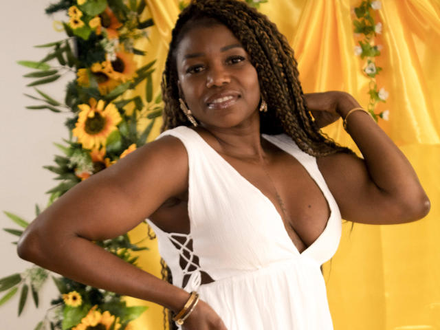 LeleFw, dark-skinned woman with long braids, wearing a deep V white dress, showing her tits, smiling, hand on head, yellow curtain, sunflowers, green leaves in background.