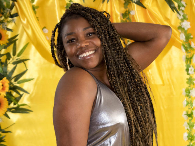 LeleFw with dark skin, long braided hair, smiling, wearing a shiny silver tank top, hand behind head, yellow backdrop with flowers. Boobs not visible, ass not visible, pussy not visible.