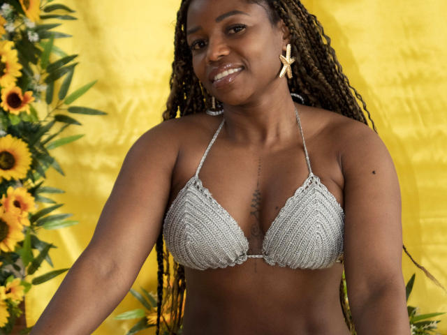 LeleFw with dark skin, long braids, small silver crocheted bikini top, showing her tits, yellow background, sunflower, and green leaves on the left, wearing star earrings, smiling.