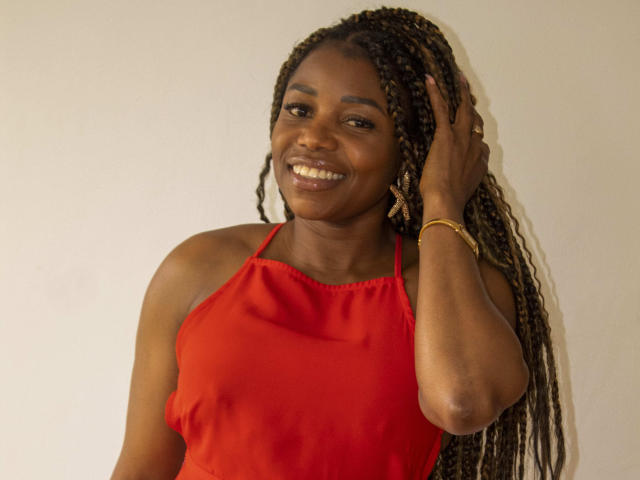 LeleFw with dark skin, long braided hair, wearing a red halter top, smiling, right hand touching head, gold bracelet, white wall background.