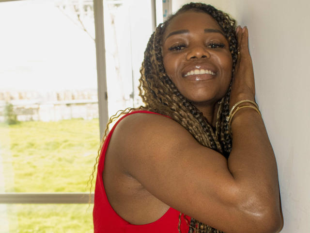 LeleFw has dark brown skin, curly braided hair, red sleeveless top, smiling, hand on white wall, gold bracelets, shiny teeth, and a relaxed, confident expression in a bright room.