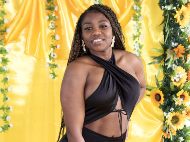 LeleFw with dark skin, long braids, wearing a black halter dress showing her tits and ass, smiling, yellow floral background, sunflowers, white flowers, and greenery.