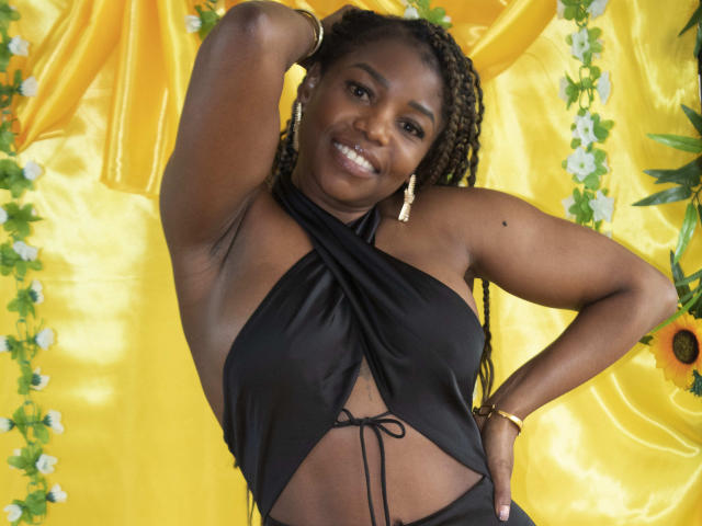 LeleFw with dark skin, smiling, wearing a black halter top showing her tits, braided hair, gold earrings, and yellow backdrop. Her right arm's raised, hand on her head.
