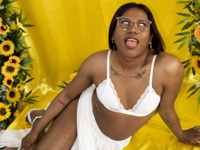 KimmySolange with dark skin, glasses, and tongue out, wearing white lace bra and skirt, sitting among sunflowers, has tattoos on chest and arm, and a cute, sexy look.