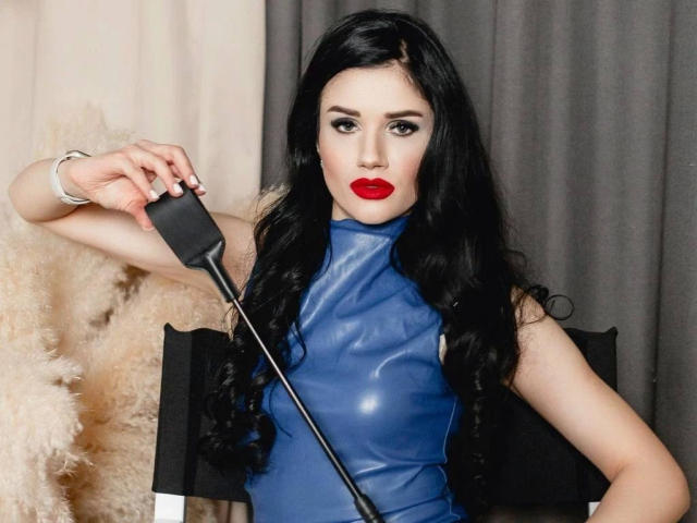 KristiTopxx – sinnlich, mit langen schwarzen Haaren, rotem Lippenstift und einem blauen Lateikleid, hält sie mit ihrer rechten Hand einen schwarzen Peitsche. Sie wirkt intensiv. Ihre Brüste sind unter dem Kleid sichtbar..