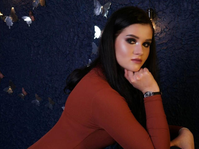 KristiTopxx-hot with long dark hair, wearing a red long-sleeve top, resting her chin on her hand, looking over her shoulder with a slight smile. Her ass is visible.