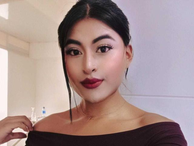 MichelleDenise has dark hair, brown eyes, and red lipstick. She's wearing an off-shoulder maroon top, a gold necklace, and a subtle smile. Her ass is not visible.