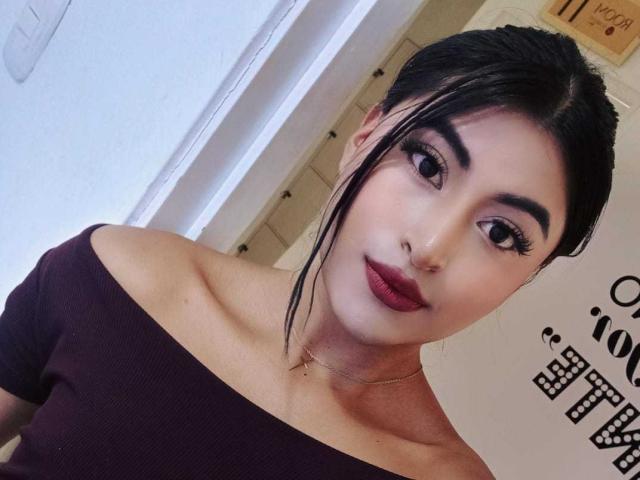 MichelleDenise has dark hair, thick eyebrows, red lipstick, and is wearing an off-shoulder purple top, showing her bare shoulders and collarbone. She's looking at the camera with a slight smile.