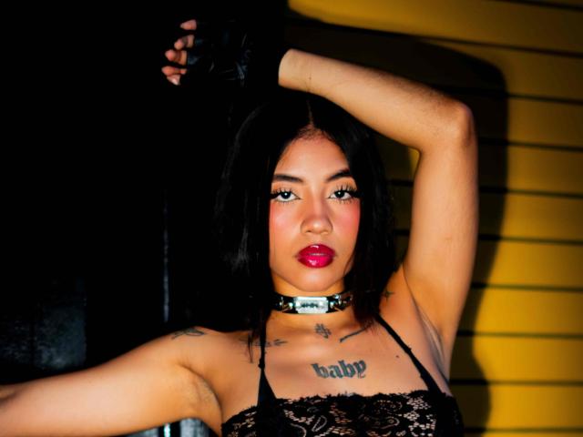 AvaRouge has black hair, red lipstick, black lace top, black choker, and black glove. She's raising her arm, showing tattoos on her chest and arm.