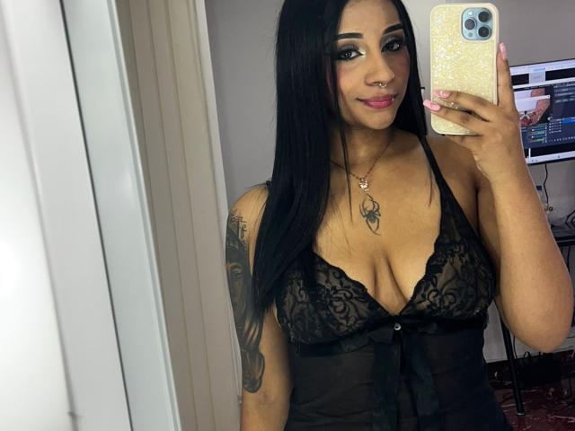 Camille69 con lunghi capelli neri, lingerie in pizzo nero, che mostra il seno, tatuaggio sul braccio destro, che tiene un telefono, sorridente, rossetto rosa, piercing al naso, in un selfie allo specchio..