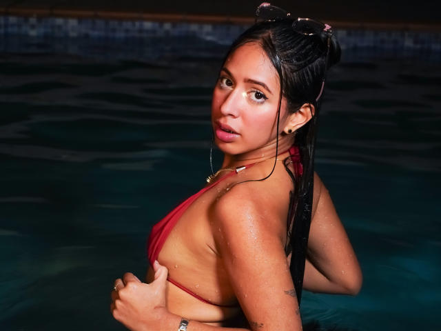 Ciamsth with wet black hair in a red bikini, covering her tits with one hand, looking over her shoulder, water droplets on her tan skin, pool at night.