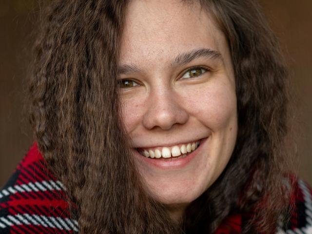 KalliFaye's smiling face with curly brown hair, green eyes, and white teeth, wearing a red and black plaid shirt. Her nose is slightly freckled.