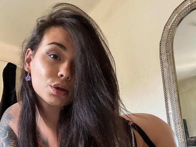 KassiKassou has long dark hair covering one eye, a nose ring, and wears black lingerie with chain straps. Tattoo on her shoulder, purple earrings, and slightly parted lips.