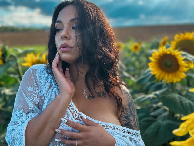 KassiKassou with wavy brown hair, white lace top, tattoos, and nose ring, touches her cheek, sunflowers in the background, under a cloudy sky, hands on her tits.