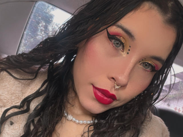 SofiBrandford has wet black hair, red lipstick, and yellow eyeshadow with black dots. She's wearing a pearl necklace and a beige fur jacket, with a nose ring.