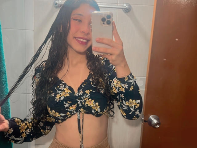 SofiBrandford with wet curly hair, wearing a dark floral bra that highlights her small tits, holding a phone in a bathroom mirror selfie, smiling.