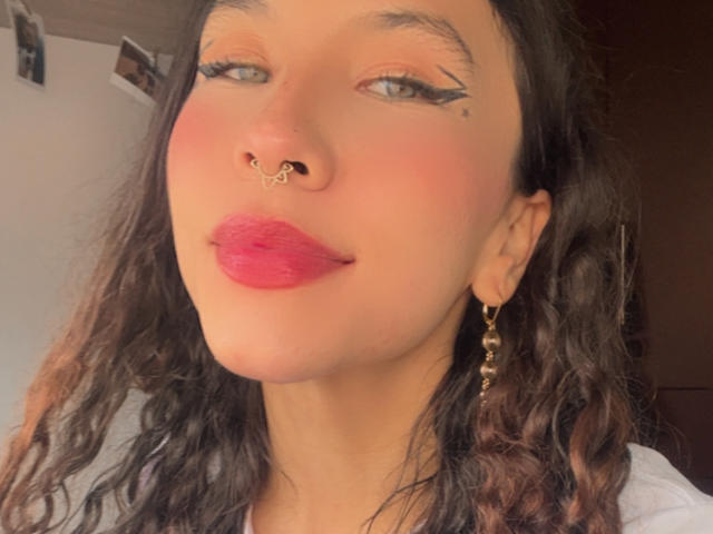 SofiBrandford with curly brown hair, nose ring, bold black eyeliner, pink lipstick, and gold earrings, looking slightly up with a subtle smile. Her skin is light and she's cute.