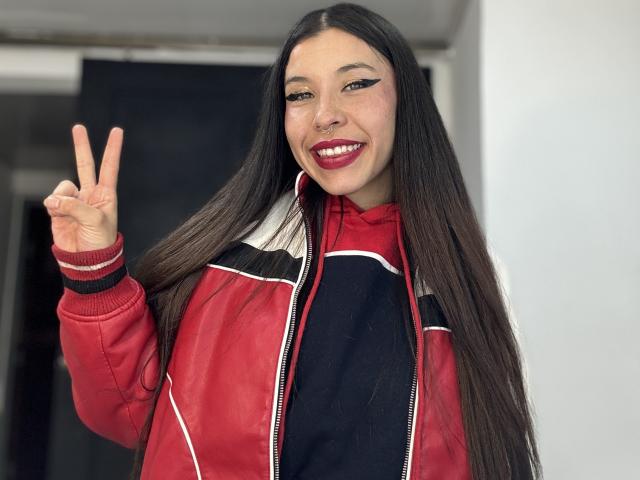 SofiBrandford with long black hair, red jacket, black shirt, nose ring, and red lipstick, flashing a peace sign, smiling with red lipstick, and black eyeliner.