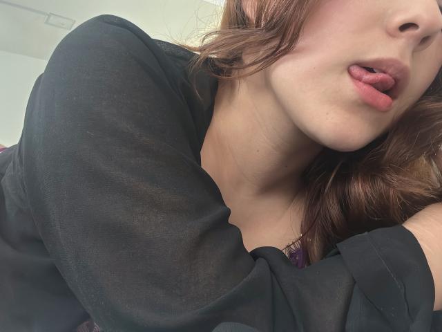 AmberlStha's face, tongue out, slightly parted lips, wearing a black sheer top, brown wavy hair, fair skin, subtle makeup, visible collarbone, upper chest, soft light, close-up shot.