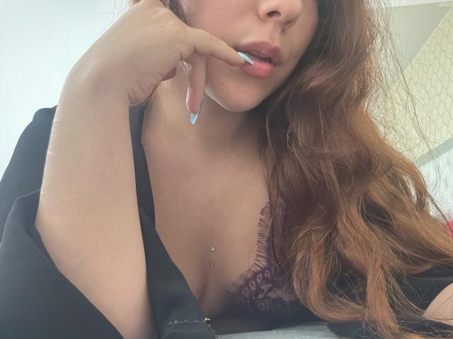 AmberlStha, com cabelo ruivo, mordendo uma unha com ponta azul, vestindo uma túnica preta, mostrando o colo com um sutiã de renda roxa, e com um piercing no mamilo, olhando para baixo..