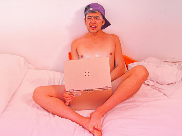 TonyDan, sin camisa, usando gafas y una gorra púrpura puesta al revés, sentado en una cama con las piernas separadas, sosteniendo una computadora portátil, con un trasero visible, húmedo y brillante..