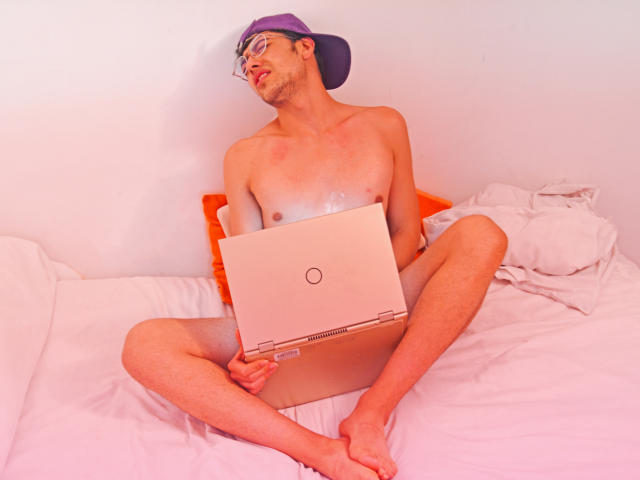 TonyDan è senza camicia, indossa un cappello viola e occhiali, è seduto su un letto con le gambe divaricate, tiene un portatile, guarda verso l'alto con un'espressione rilassata, con i glutei scoperti..