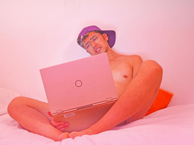 TonyDan, nu, usando um boné roxo e óculos, está sentado em uma cama com um laptop, com as pernas abertas, mostrando sua bunda nua e um pênis flácido levemente visível..