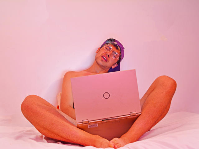 TonyDan zit zonder shirt, met gespreide benen, en houdt een roze laptop op zijn schoot. Hij draagt een paarse hoofdband en een bril, en ziet er moe uit met een licht geopende mond..