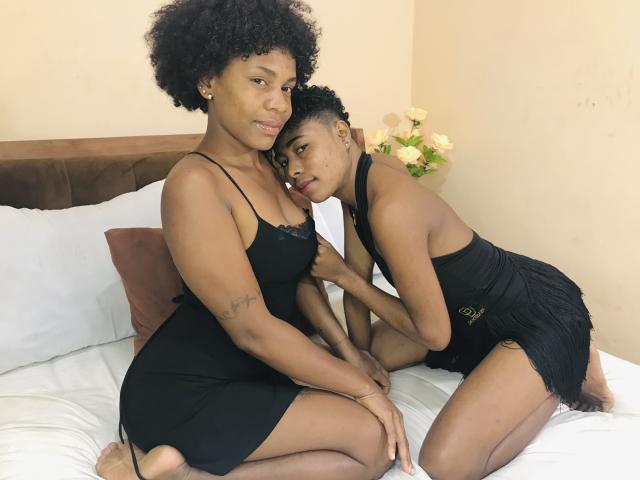 JulianaSilvie with curly black hair, wearing a black dress, sits on a bed with her arm around a shorter JulianaSilvie in a black fringed dress, showing her small tits.