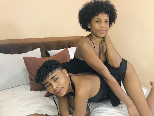 JulianaSilvie with curly black hair, small tits, and a tattoo on her arm, wearing a black halter top, sitting on a bed with white and brown pillows.