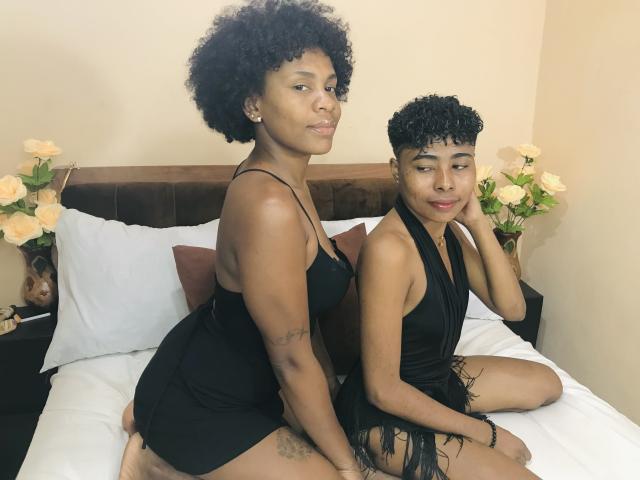Two black women with curly hair, wearing black dresses, sitting on a bed. JulianaSilvie on the left with a tattoo on her thigh, JulianaSilvie on the right, looking back.