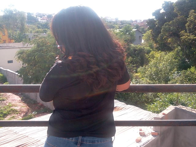 MariahSquirting69, con capelli castani ondulati, felpa nera e jeans blu, in piedi con la schiena rivolta verso la telecamera, luce solare tra i capelli, appoggiata a una ringhiera di metallo, ambientazione esterna, con vegetazione visibile..