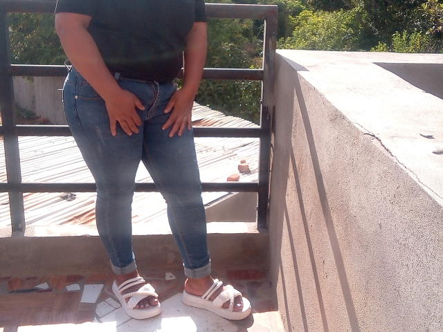 MariahSquirting69's hands on her blue jeans, black top, white strappy sandals, standing on sunlit balcony, concrete wall to right, metal railing behind, greenery in background.