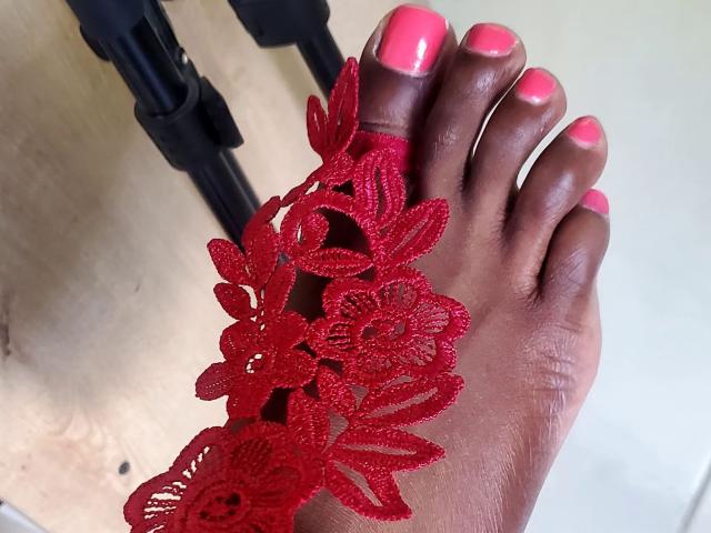 ChelseyeBonny's foot with dark brown skin, pink nail polish, and a red lace flower design covering the toes. Black chair legs in the background.
