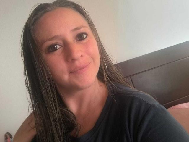 CelesteStorm has wet brown hair, a slight smile, and brown eyes, wearing a black shirt, with her tits barely visible, in a bedroom with a dark wooden headboard.