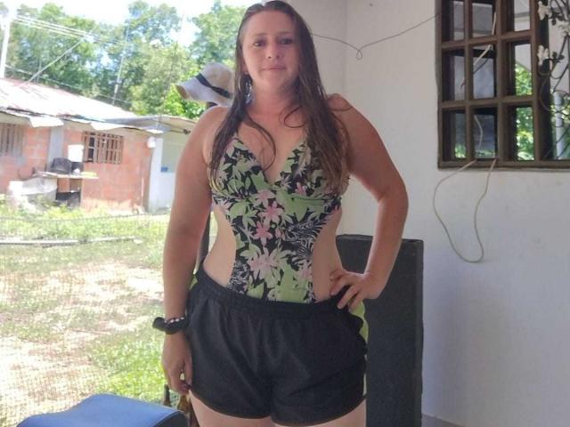 CelesteStorm with wet brown hair, wearing a floral one-piece and black shorts, stands with hands on hips, showing her tits in a sunlit, outdoor setting.