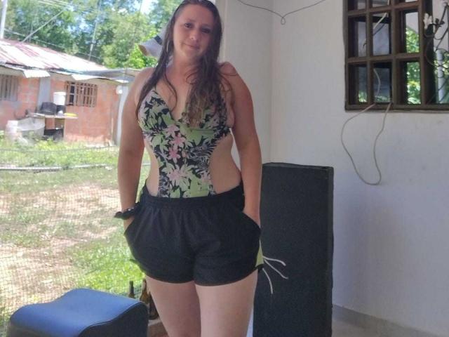 CelesteStorm with long brown hair, wearing a floral one-piece and black shorts, standing with hands in pockets, showing her ass and a sideboob.