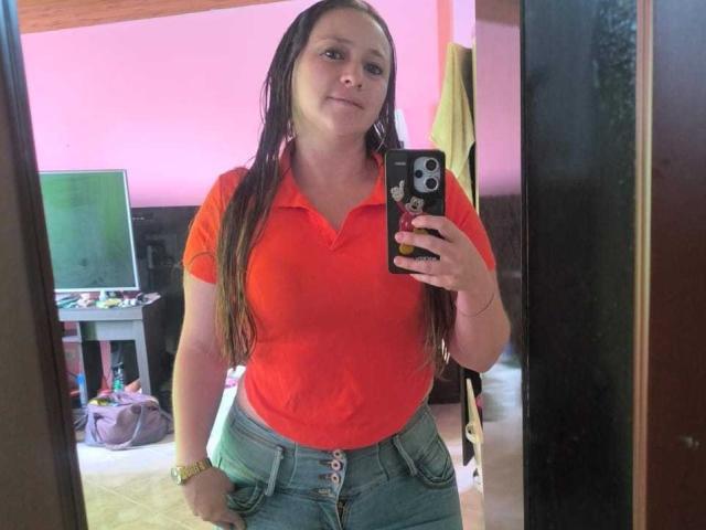 CelesteStorm with long brown hair, red polo, and blue jeans, holding phone in mirror selfie, smiling, showing her small tits, and wearing a gold bracelet.