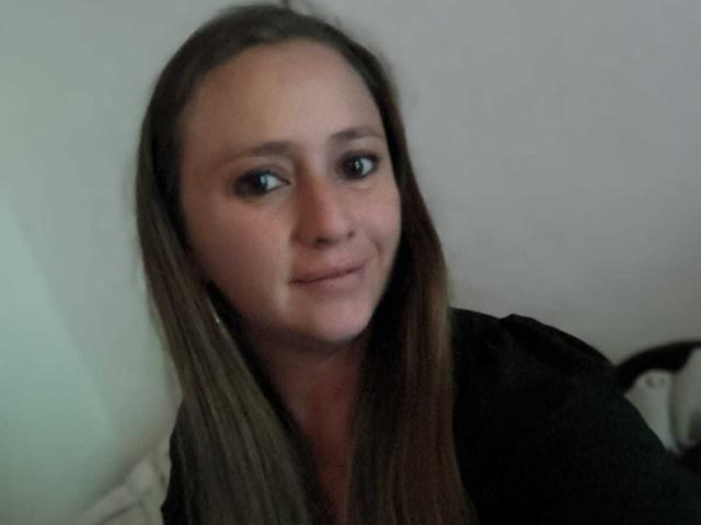 CelesteStorm has straight brown hair, a slight smile, and is wearing a black top, showing her pale skin and brown eyes. She's sitting in a relaxed, indoor setting.