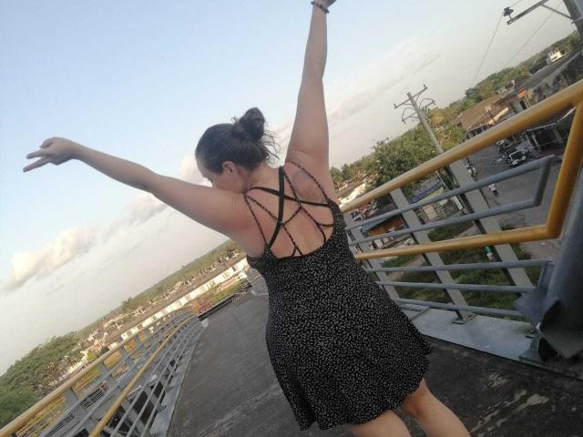CelesteStorm has her arms raised, wearing a black dress with a crisscross back, standing on a bridge, facing away, with a city and water in the background.