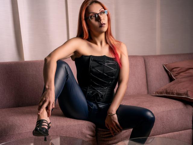 ArtemisFlyve with red hair, glasses, black strapless corset, blue leather pants, sitting on pink sofa, legs spread, black heels, looking up, hands on knee, confident, sexy.