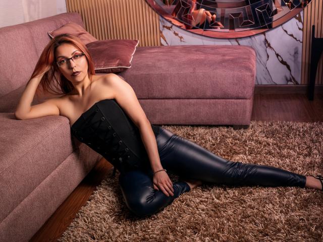 ArtemisFlyve with red hair, glasses, black strapless corset, and black leather pants, sitting on a shaggy rug, hand on pink sofa, looking at camera. Boobs, ass visible.