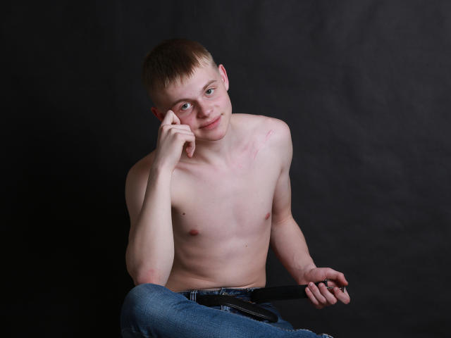 Shirtless HarleyQuinXO with a slim, pale body and small nipples, sitting with a black belt in his right hand, short brown hair, and a slight smile, against a dark background.