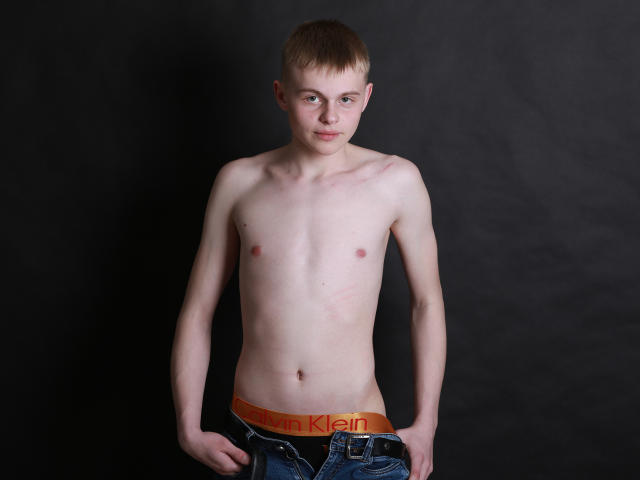 HarleyQuinXO, shirtless, skinny with small tits, pale skin, short brown hair, looking at camera, wearing unbuttoned jeans with orange Calvin Klein waistband, standing against dark background.